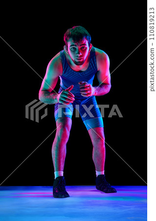 Sportsman, freestyle wrestler in blue sport tights stands in attack position and looking ta camera against black background in mixed neon lights. Sportsman, freestyle wrestler in blue sport tights stands in attack position and looking ta camera against black background in mixed neon lights. 110819213