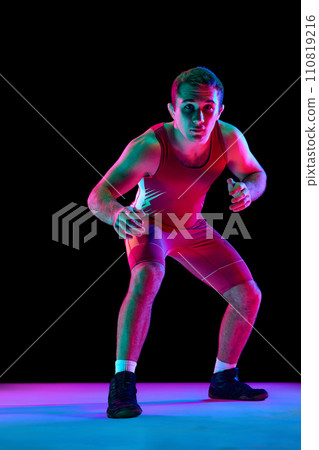 Young man, professional freestyle fighter stands in attacking pose and looking at camera against black background in mixed neon lights. 110819216
