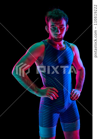 Focused and competitive freestyle wrestler in modern blue sportwear put his hands on hips and looking at camera against black background in mixed neon lights. 110819222