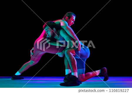 Two young, skilled and professional freestyle wrestlers in red and blue fighting uniform against black background in mixed neon lights. Two young, skilled and professional freestyle wrestlers in red and blue fighting uniform against black background in mixed neon lights. 110819227