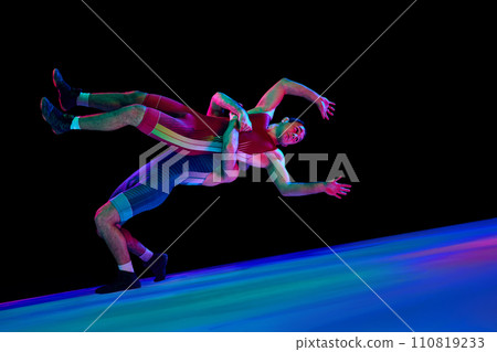 Wrestler in blue uniform lifts opponent in red, both in dynamic bridge position against black background in mixed neon lights. Mixed martial art. 110819233
