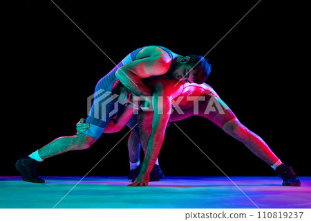 Two strong wrestlers in blue and red wrestling uniform fighting and making hip throw in motion against black background in mixed neon lights. Two strong wrestlers in blue and red wrestling uniform fighting and making hip throw in motion against black background in mixed neon lights. 110819237