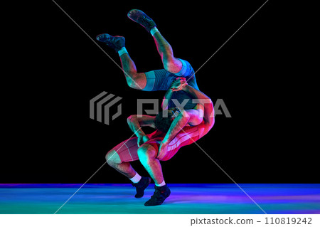 Two young men in blue and red wrestling tights wrestling and making suplex fight against black background in mixed neon lights. 110819242