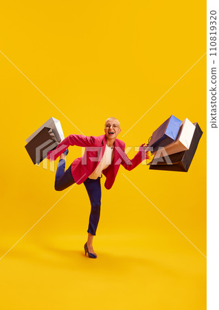 Happy, delightful woman running with running with large bags after productive shopping against vivid yellow studio background. 110819320