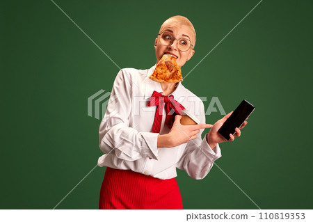 Confident businesswoman with pizza slice holding in one hand coffee and pointing to smartphone with blank screen against green studio background. 110819353