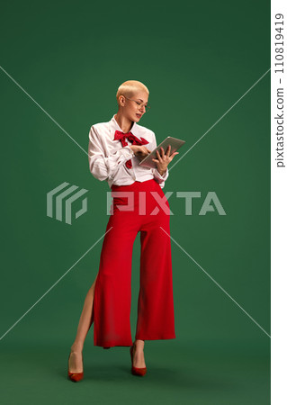 Confident business lady in trendy bright clothes working online using tablet against green studio background. 110819419