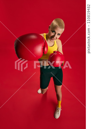 Top view portrait of confident female boxer in yellow top, black shorts, red gloves boxing looking to camera against red background. Top view portrait of confident female boxer in yellow top, black shorts, red gloves boxing looking to camera against red background. 110819482