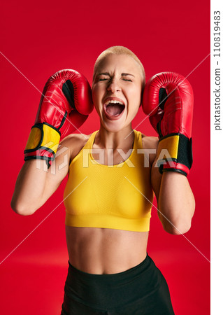 Young sporty emotional woman in sport wear and boxing gloves shouting loudly and closed eyes and ears of her self noise against red studio background. Young sporty emotional woman in sport wear and boxing gloves shouting loudly and closed eyes and ears of her self noise against red studio background. 110819483