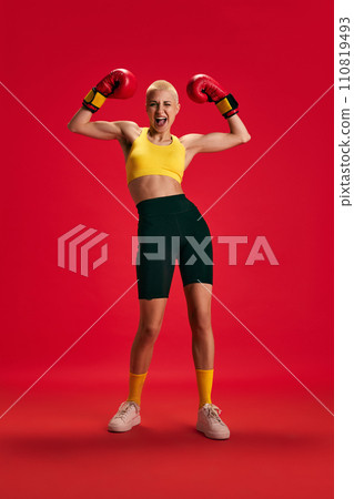 Full-length portrait of young emotional woman in sportwear and boxing gloves posing against bright red background. Full-length portrait of young emotional woman in sportwear and boxing gloves posing against bright red background. 110819493