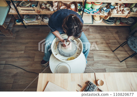 A woman ceramist at a pottery wheel in a workshop. The beginning of shaping a white clay product on a potter's wheel A woman ceramist at a pottery wheel in a workshop. The beginning of shaping a white clay product on a potter's wheel 110820677