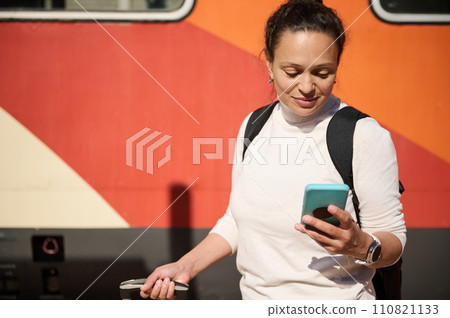 Happy tourist woman using mobile phone while waiting on station platform on the background of a colorful electric train Happy tourist woman using mobile phone while waiting on station platform on the background of a colorful electric train 110821133