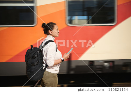 Side portrait woman commuter with suitcase, holding mobile phone, standing at railway platform while travelling 110821134