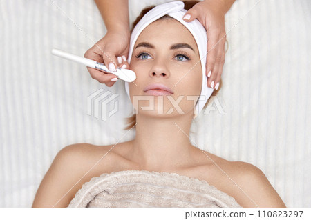 female hand touching the patient's face with a brush. beautician applies the mask to female face. facial skin care. top view female hand touching the patient's face with a brush. beautician applies the mask to female face. facial skin care. top view 110823297