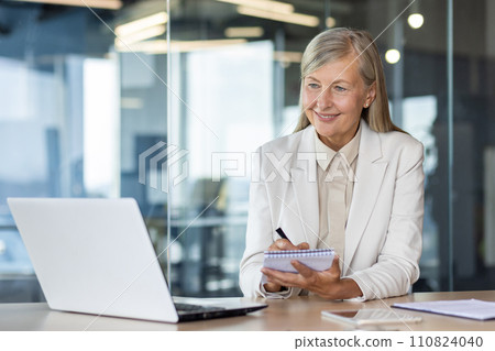 Smiling senior business woman sitting in the office at the table in front of the notebook, looking at the screen and writing down information and data in the notebook. 110824040