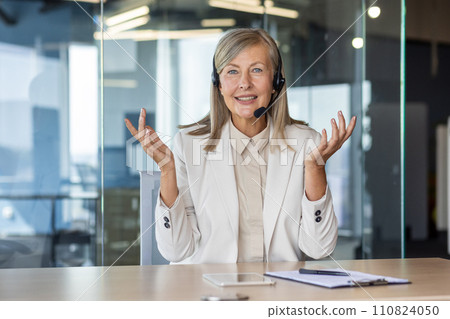 Portrait of a smiling gray-haired senior woman sitting in the office at a desk in front of a camera in a headset and talking on a video call, gesturing with her hands 110824050