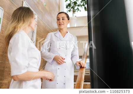Skilled female doctor and young colleague nurse woman talking standing on staircase in medic clinic, smiling, discussing medical practice, enjoying networking, job communication. Concept of healthcare 110825373