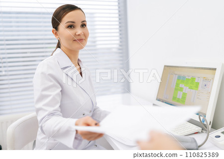Smiling female doctor giving prescription paper to unrecognizable patient at checkup appointment sitting at table. Physician handing out document for treatment and medicine against disease. 110825389