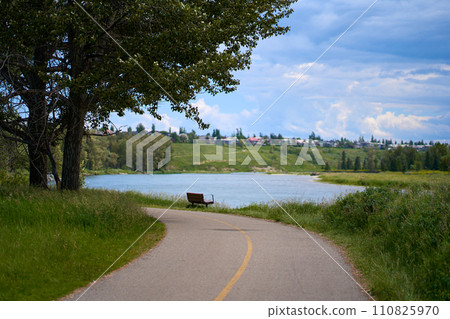 A path in a beautiful summer city park by the river. A bench on the shore of a calm lake in the forest. 110825970