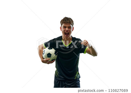 Portrait of emotional young man in black uniform, soccer player with ball emotionally shouting isolated over white background with grass flooring 110827089