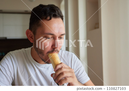 Closeup portrait of hungry man eating delicious sandwich at home on background of kitchen. Handsome male enjoying vegetarian dinner indoors. Caucasian guy eating toast in morning, slow motion. 110827103