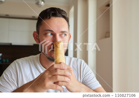 Closeup portrait of hungry man eating delicious sandwich at home on background of kitchen. Handsome male enjoying vegetarian dinner indoors. Caucasian guy eating toast in morning, slow motion. 110827106