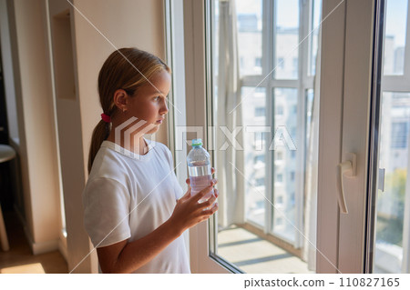 Back view of thirsty little 10s girl drinking water from plastic bottle, break and workout with hydration standing by window at home in sunny day. Kid drinks cup of fresh pure filtered mineral water. 110827165