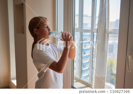 Back view of thirsty little 10s girl drinking water from plastic bottle, break and workout with hydration standing by window at home in sunny day. Kid drinks cup of fresh pure filtered mineral water. 110827166