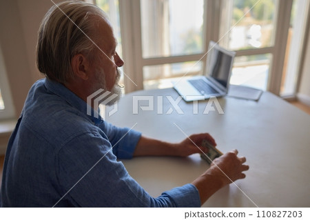 Portrait of bearded senior pensioner male with wrinkled hands counting Russian ruble banknotes sitting at home table. 110827203