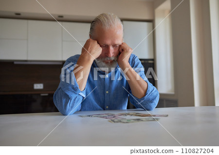 Portrait of bearded senior pensioner male with wrinkled hands counting Russian ruble banknotes sitting at home table. 110827204