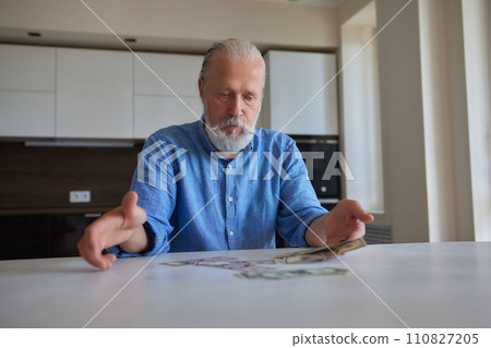 Portrait of bearded senior pensioner male with wrinkled hands counting Russian ruble banknotes sitting at home table. 110827205