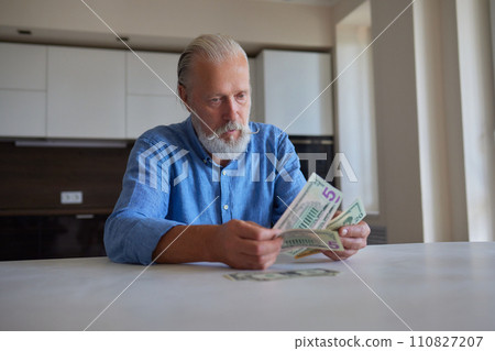 Portrait of bearded senior pensioner male with wrinkled hands counting Russian ruble banknotes sitting at home table. 110827207