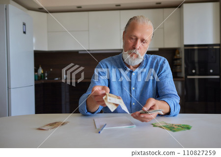 Portrait of bearded senior pensioner male with wrinkled hands counting Russian ruble banknotes sitting at home table. 110827259