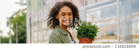 Smiling woman florist holding flower pot while standing on greenhouse yard background Smiling woman florist holding flower pot while standing on greenhouse yard background 110828009
