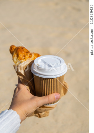 Tasty fresh croissants with coffee in paper cup in female hands on beach sea background. Breakfast outdoors wellbeing concept. FPV. Take away food Tasty fresh croissants with coffee in paper cup in female hands on beach sea background. Breakfast outdoors wellbeing concept. FPV. Take away food 110829423