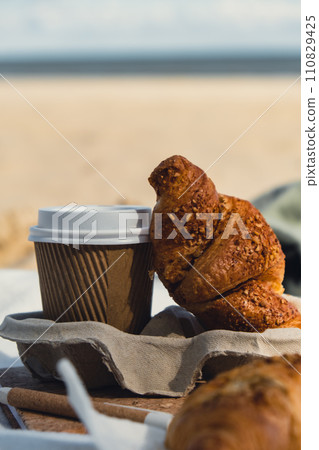 Tasty fresh croissants with coffee in paper cup on sandy beach sea coast background. Breakfast outdoors wellbeing concept. Take away food. Coffee alternatives Tasty fresh croissants with coffee in paper cup on sandy beach sea coast background. Breakfast outdoors wellbeing concept. Take away food. Coffee alternatives 110829425