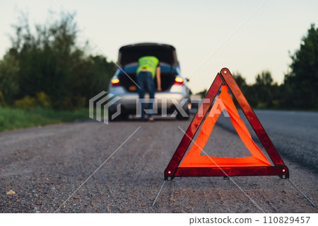 Unrecognizable sad driver in reflective vest. Male driver standing near broken car with open up hood. Red triangle to warn other road users of car breakdown or engine failure stop at countryside Unrecognizable sad driver in reflective vest. Male driver standing near broken car with open up hood. Red triangle to warn other road users of car breakdown or engine failure stop at countryside 110829457