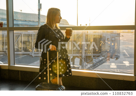 A young woman, holding a cup of coffee is ready for boarding with her suitcase. A young woman, holding a cup of coffee is ready for boarding with her suitcase. 110832763