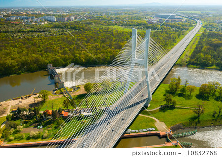 Cars drive on cable-stayed Redzinski Bridge over river flowing near scenic Wroclaw. Pylon bridge surrounded by lush green forests aerial motion along bridge 110832768