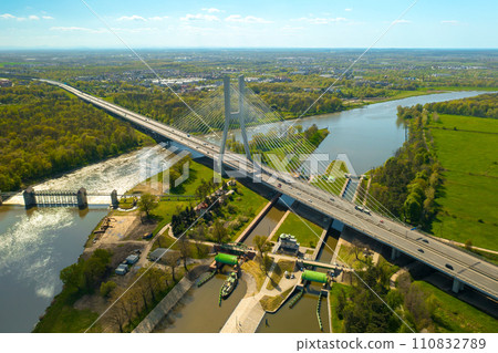 Vehicles drive on massive Redzinski Bridge in Poland on sunny spring day. Powerful cable-stayed pylon bridge built over Oder river aerial view 110832789