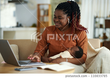 Portrait of smiling Black woman with sleeping baby having online meeting with colleagues 110833785