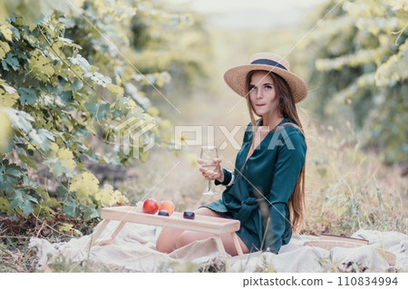 Woman picnic vineyard. Happy woman with a glass of wine at a picnic in the vineyard, wine tasting at sunset and open nature in the summer. Romantic dinner, fruit and wine. 110834994