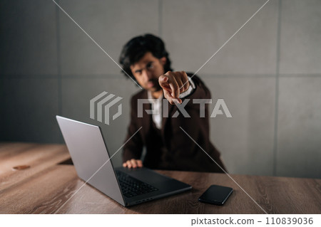 Portrait of serious successful businessman in suit looking at camera and pointing finger forward calling to action. Front view of business man working on laptop sitting at table modern office. 110839036