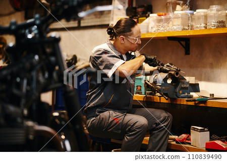 Repairman in protective googles filing motorcycle detail at his workbench Repairman in protective googles filing motorcycle detail at his workbench 110839490