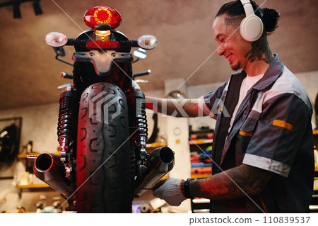 Smiling mechanic listening to music in headphones when repairing motorcycle Smiling mechanic listening to music in headphones when repairing motorcycle 110839537