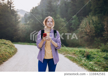Girl tourist walking in forest and using smartphone. Young female with backpack hiking on the road in nature. Beautiful 30s woman walking on nature in sport clothes. Girl tourist walking in forest and using smartphone. Young female with backpack hiking on the road in nature. Beautiful 30s woman walking on nature in sport clothes. 110842581
