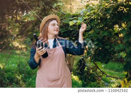 30s Woman in hat tasting red wine in vineyard. Portrait of pretty young woman holding bottle of wine on sunny day. Natural wine industry. Mockup for design 110842582