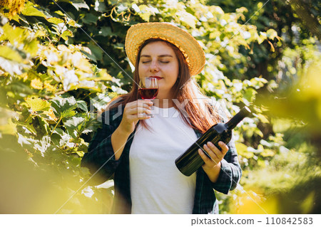30s Woman in hat tasting red wine in vineyard. Portrait of pretty young woman holding glass of wine and bottle of wine. Mockup for design, Natural wine industry. 110842583