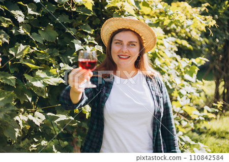 30s Woman in hat tasting red wine in vineyard. Portrait of pretty young woman holding glass of wine. Happy vintner drinks wine after successful grape harvesting, Natural wine industry. 30s Woman in hat tasting red wine in vineyard. Portrait of pretty young woman holding glass of wine. Happy vintner drinks wine after successful grape harvesting, Natural wine industry. 110842584