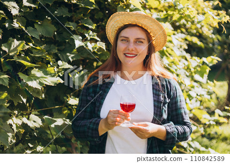 30s Woman in hat tasting red wine in vineyard. Portrait of pretty young woman holding glass of wine. Happy vintner drinks wine after successful grape harvesting, Natural wine industry. 110842589