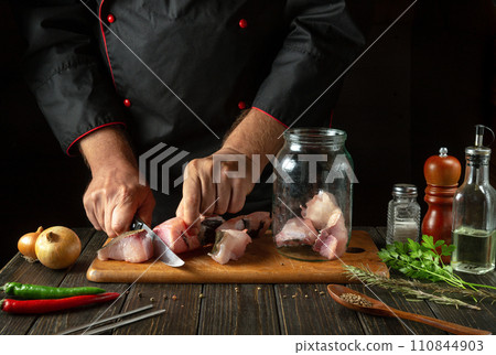 The chef cuts carp fish to prepare herring in a jar with spices. The concept of salting fish in a restaurant or hotel kitchen The chef cuts carp fish to prepare herring in a jar with spices. The concept of salting fish in a restaurant or hotel kitchen 110844903